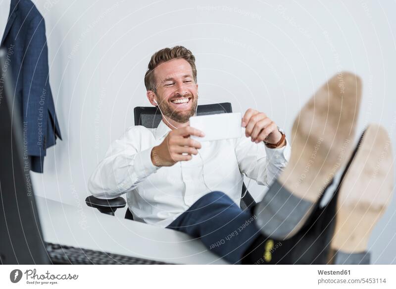 Geschäftsmann mit Kopfhörern sitzt am Schreibtisch mit erhobenen Füßen und schaut auf sein Handy Businessmann Businessmänner Geschäftsmänner Portrait Porträts