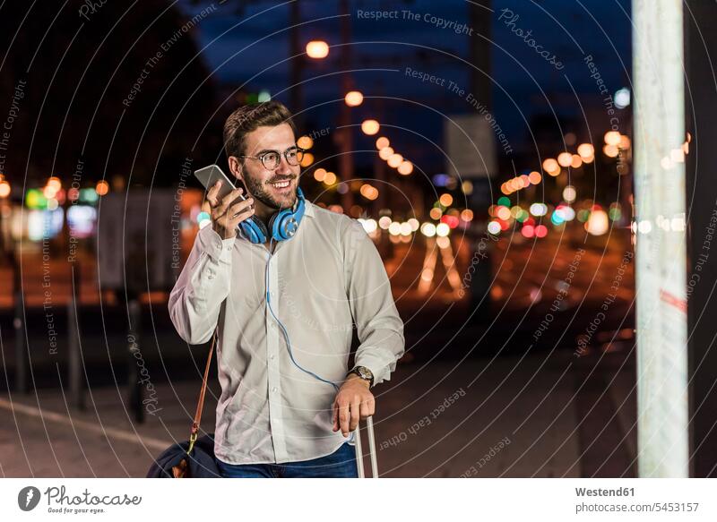 Junger Mann in der Stadt mit Kopfhörern und Handy in der Nacht Männer männlich Kopfhoerer staedtisch städtisch lächeln Mobiltelefon Handies Handys Mobiltelefone
