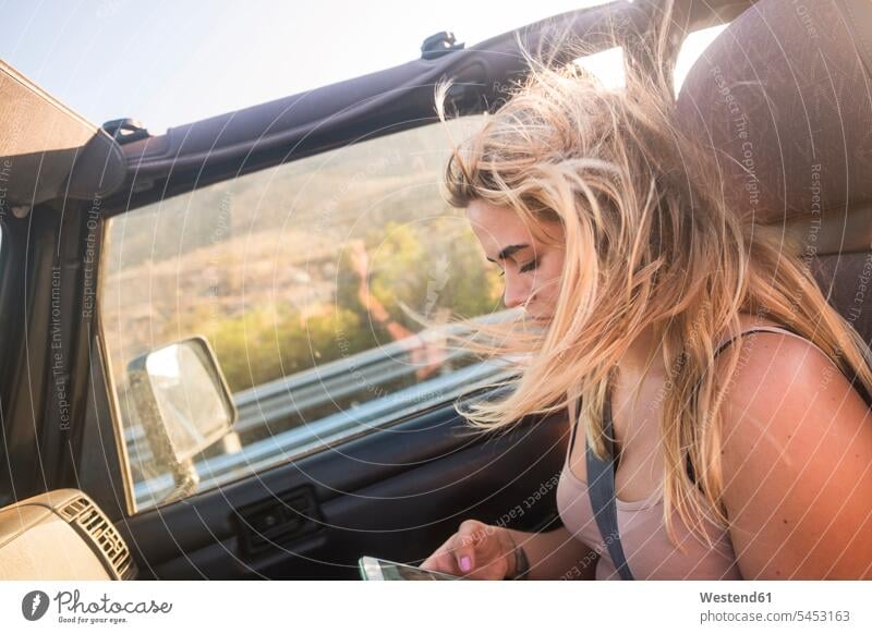 Blonde Frau mit wehenden Haaren sitzt im Auto und telefoniert benutzen benützen Smartphone iPhone Smartphones wehende Haare wehendes Haar sitzen sitzend blond
