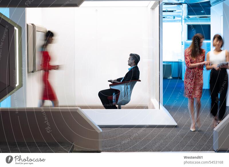 Geschäftsmann sitzt auf einem Stuhl in einer belebten Büroetage und arbeitet Office Büros futuristisch Zukunft Future Visionär Geschäftigkeit geschäftig