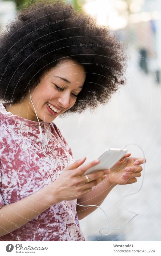 Porträt einer lächelnden jungen Frau mit Kopfhörern, die auf ein Mobiltelefon schaut weiblich Frauen Smartphone iPhone Smartphones Erwachsener erwachsen Mensch