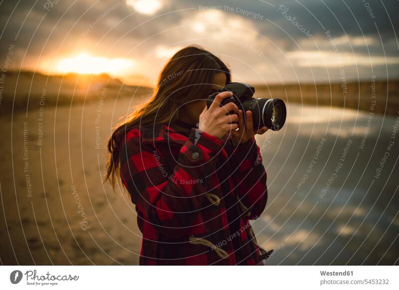 Junge Frau, die bei Sonnenuntergang am Strand mit der Kamera fotografiert weiblich Frauen fotografieren Kameras Erwachsener erwachsen Mensch Menschen Leute