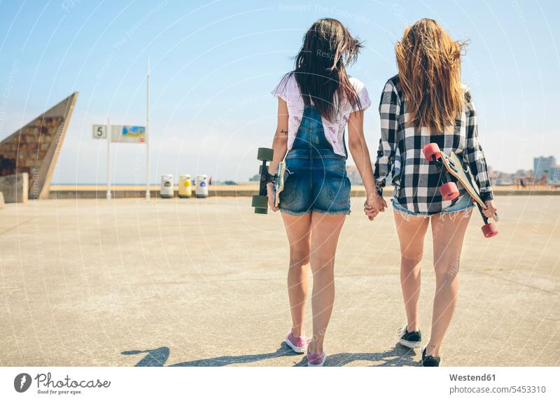 Rückenansicht von zwei jungen Frauen mit Longboards, die Hand in Hand auf der Strandpromenade gehen gehend geht Sexy Rückansicht Rueckansicht Rueckenansicht