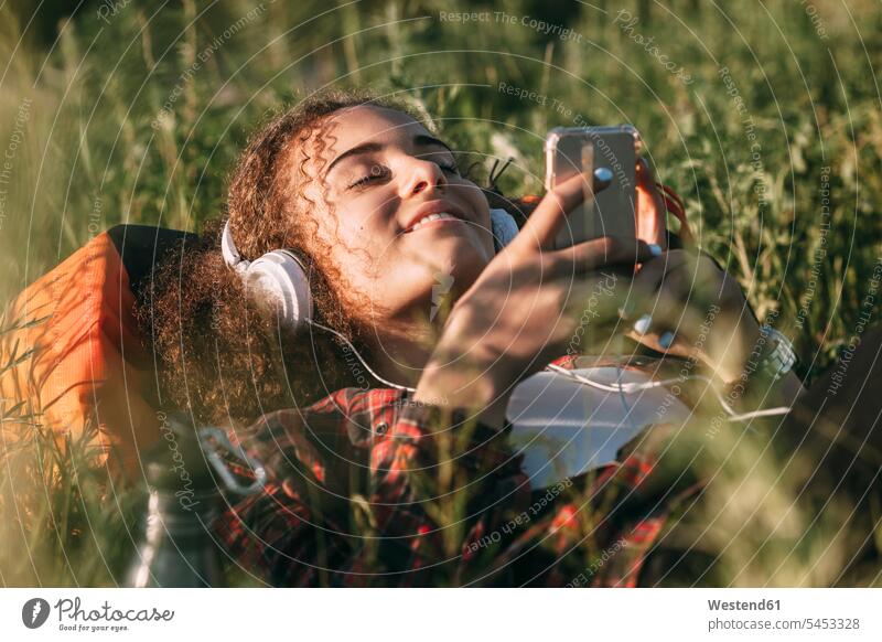 Teenagerin mit Rucksack auf einer Wiese liegend und mit Kopfhörer und Handy Musik hören junges Mädchen Teenagerinnen weiblich junge Frau Kopfhoerer Jugendliche