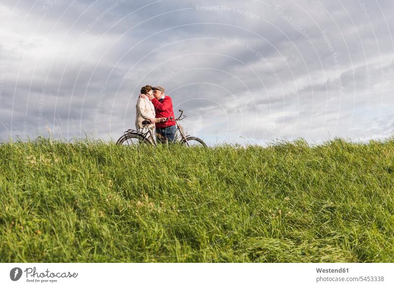 Älteres Ehepaar mit Fahrrädern küsst sich in ländlicher Landschaft unter bewölktem Himmel Paar Pärchen Paare Partnerschaft Senior ältere Männer älterer Mann