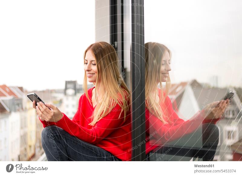 Lächelnde junge Frau sitzt am Fenster in Stadtwohnung und schaut auf Handy lächeln Mobiltelefon Handies Handys Mobiltelefone sitzen sitzend weiblich Frauen