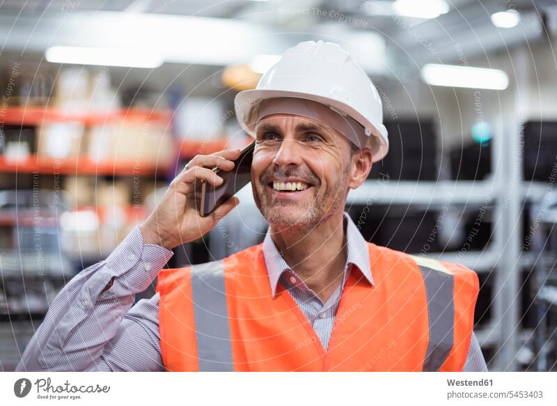 Lächelnder Mann in der Fabrikhalle mit Sicherheitsweste und Schutzhelm, der mit dem Handy telefoniert Männer männlich telefonieren anrufen Anruf telephonieren