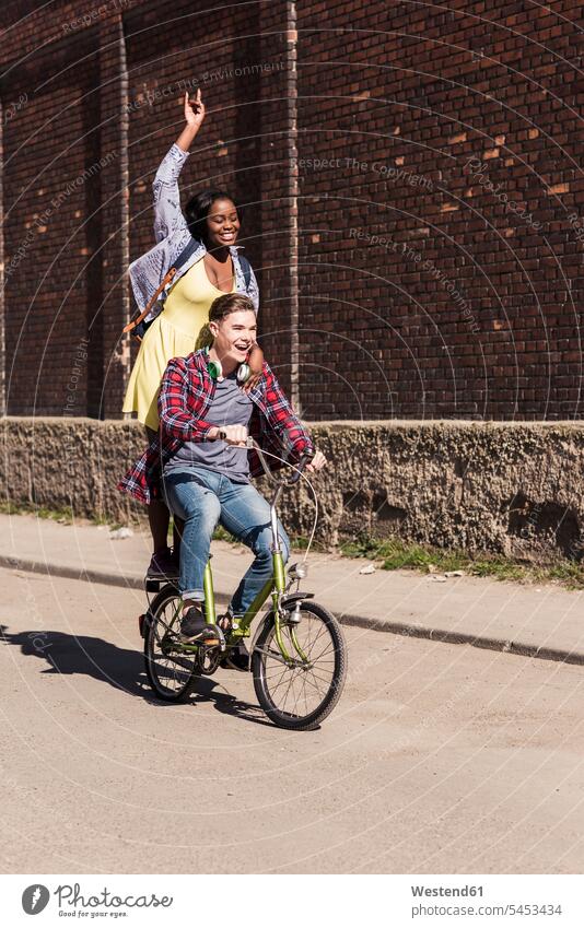 Junger Mann fährt Fahrrad mit seiner Freundin auf dem Gepäckträger stehend aktiv fröhlich Fröhlichkeit Frohsinn Heiterkeit Gemeinsam Zusammen Miteinander