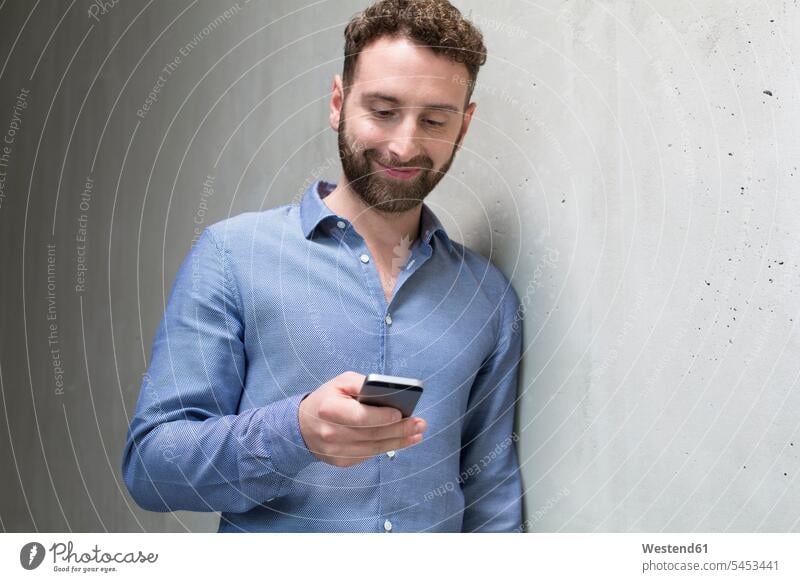 Lächelnder Mann schaut mit seinem Handy auf Betonwand Männer männlich Betonwände Betonwaende Mobiltelefon Handies Handys Mobiltelefone lächeln Erwachsener
