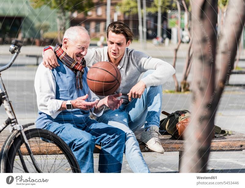 Älterer Mann und erwachsener Enkel mit Basketballgespräch auf einer Bank sitzen sitzend sitzt Enkelsöhne Enkelsohn Sitzbänke Bänke Sitzbank Großvater Opa