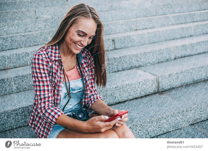 Lächelnde junge Frau sitzt auf der Treppe und schaut auf ihr Handy Mobiltelefon Handies Handys Mobiltelefone lächeln weiblich Frauen Treppenaufgang Telefon