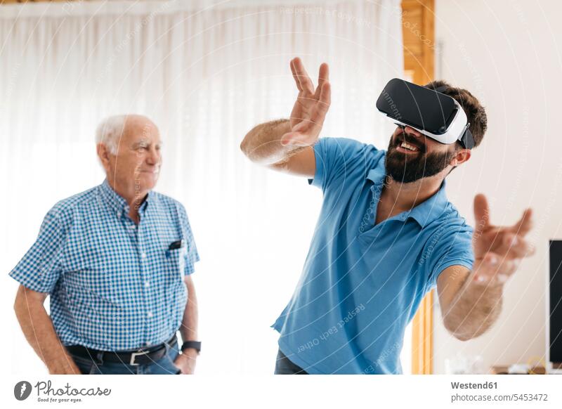 Lächelnder Mann benutzt zu Hause eine Virtual-Reality-Brille, während sein Großvater ihm zusieht Virtual Reality Brille Virtual Reality-Brille VR Brille Enkel