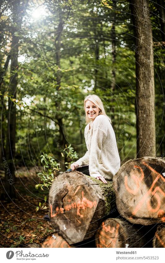 Porträt einer glücklichen Frau, die mit einer Tafel auf einem Holzstapel im Wald sitzt Forst Wälder lachen weiblich Frauen Portrait Porträts Portraits positiv