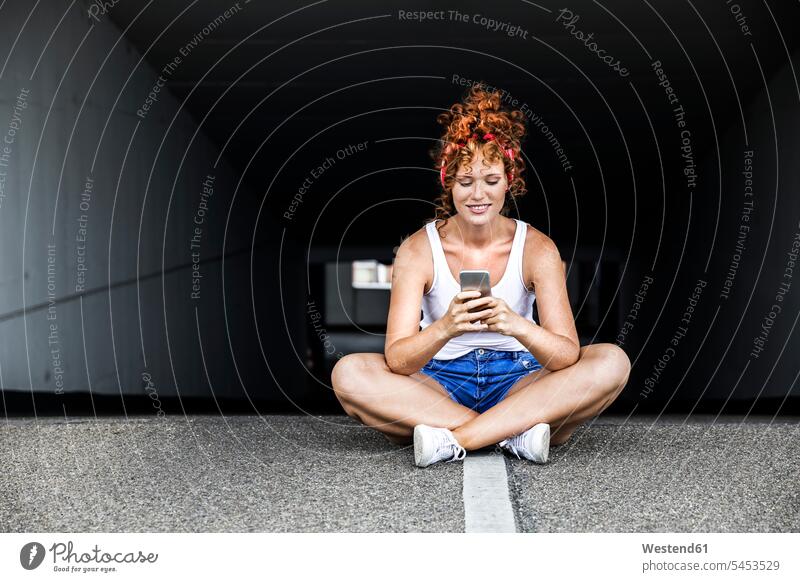 Rothaarige Frau sitzt auf Parkdeck und hält Handy weiblich Frauen Mobiltelefon Handies Handys Mobiltelefone lächeln sitzen sitzend Erwachsener erwachsen Mensch