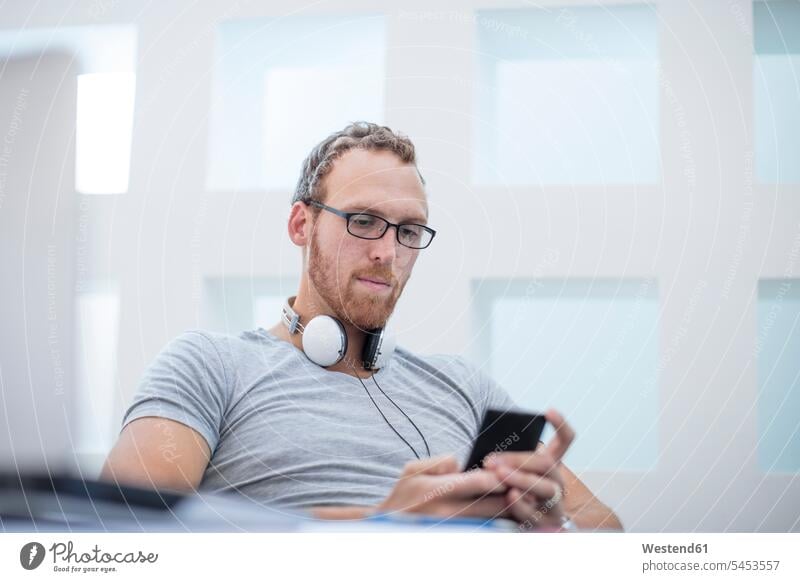 Junger Mann mit Kopfhörern um den Hals liest Textnachrichten auf seinem Telefon cool Coolness junger Mann junge Männer lesen Lektüre Büro Office Büros SMS