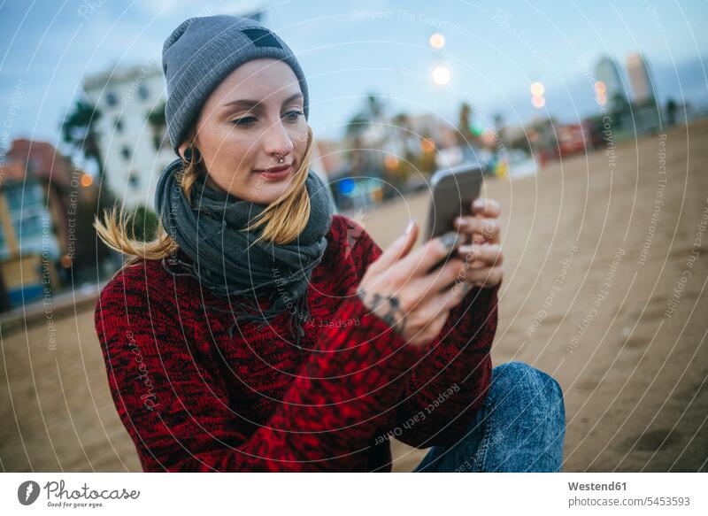 Junge Frau sitzt im Winter mit dem Handy am Strand Beach Straende Strände Beaches weiblich Frauen sitzen sitzend Mobiltelefon Handies Handys Mobiltelefone