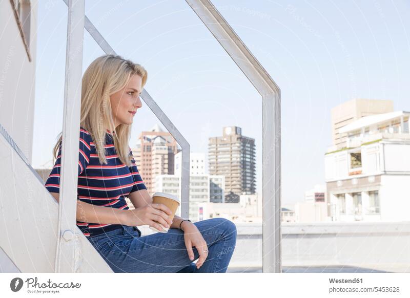 Junge Frau, die Kaffee trinkt und auf einer Treppe auf einer Dachterrasse sitzt weiblich Frauen blond blonde Haare blondes Haar Kaffeebecher Becher