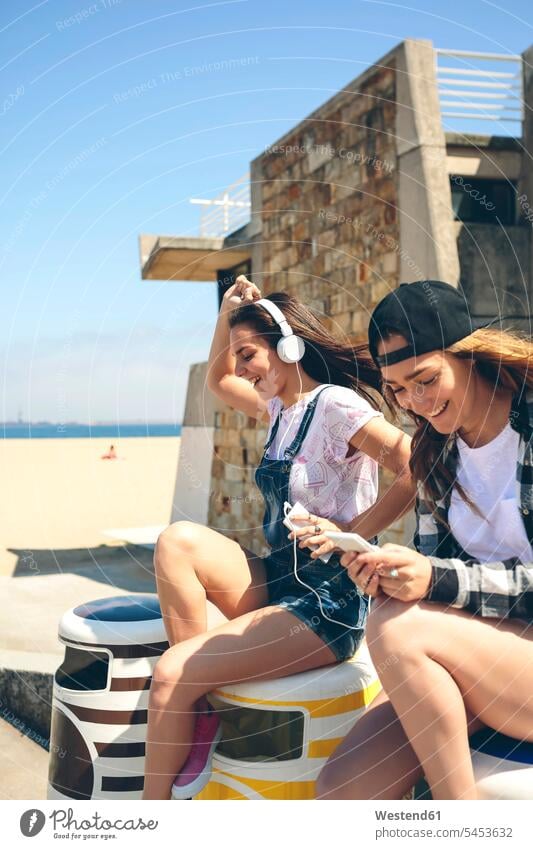 Zwei glückliche junge Frauen, die neben dem Strand Musik hören und Spaß haben Kopfhörer Kopfhoerer sitzen sitzend sitzt hoeren Smartphone iPhone Smartphones