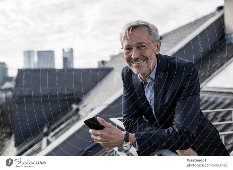 Grauhaariger Geschäftsmann auf dem Balkon hält Smartphone mit einem Lächeln in die Kamera lächeln Businessmann Businessmänner Geschäftsmänner Handy Mobiltelefon