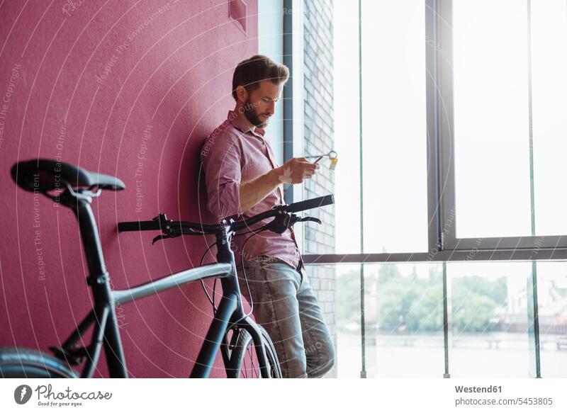 Mann mit Fahrrad steht in modernem Büro und schaut auf Handy Mobiltelefon Handies Handys Mobiltelefone Bikes Fahrräder Räder Rad Männer männlich Telefon