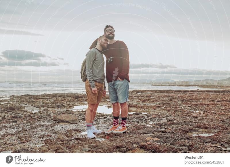 Glückliches junges schwules Paar steht am Strand Pärchen Paare Partnerschaft Beach Straende Strände Beaches Mensch Menschen Leute People Personen Homosexualität