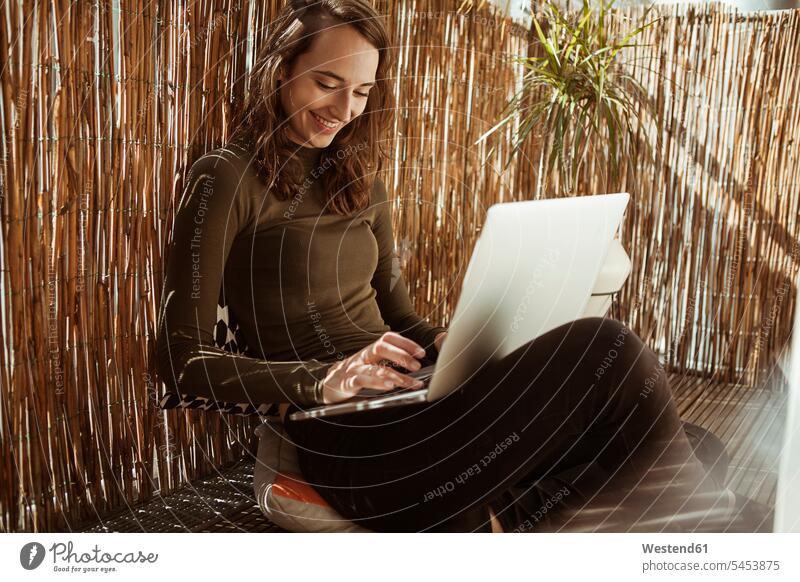 Lächelnde junge Frau sitzt mit Laptop auf dem Balkon lächeln Notebook Laptops Notebooks sitzen sitzend weiblich Frauen Computer Rechner Erwachsener erwachsen