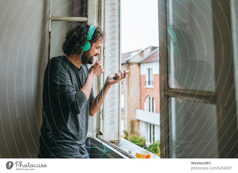 Mann telefoniert mit Handy in seinem Zimmer am Fenster Männer männlich Kopfhörer Kopfhoerer Erwachsener erwachsen Mensch Menschen Leute People Personen