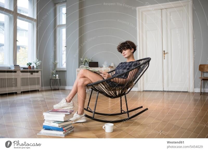 Junge Frau sitzt zu Hause auf einem Schaukelstuhl und liest ein Buch lesen Erwachsener Mensch sitzen Wohnung Wohnzimmer Arbeitszimmer Wohnen Zimmer Kaffee