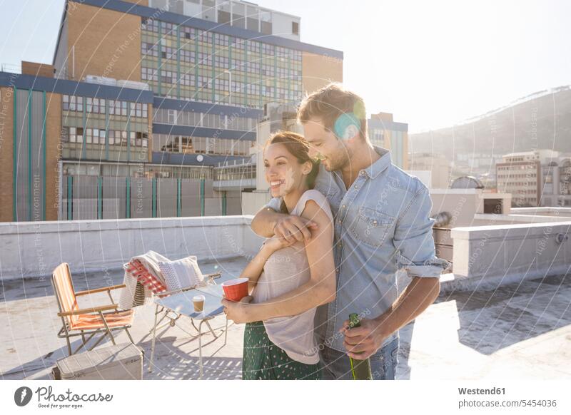 Junges Paar feiert auf einer Dachterrasse und umarmt sich bei Sonnenuntergang gemischtrassige Person Spaß Spass Späße spassig Spässe spaßig Freizeit Muße Sommer