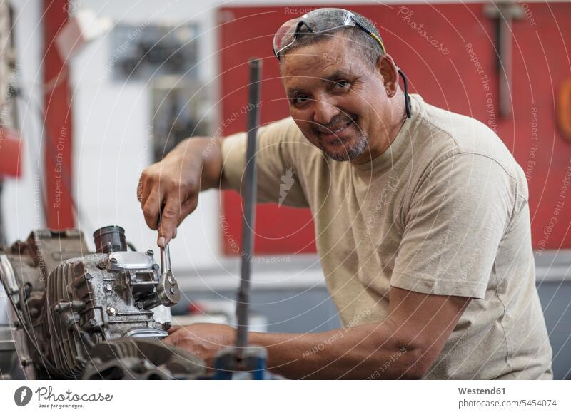 Lächelnder Mechaniker bei der Arbeit am Motorradmotor in der Werkstatt lächeln Motorräder reparieren Reparatur arbeiten Monteur Kraftfahrzeug Verkehrsmittel KFZ