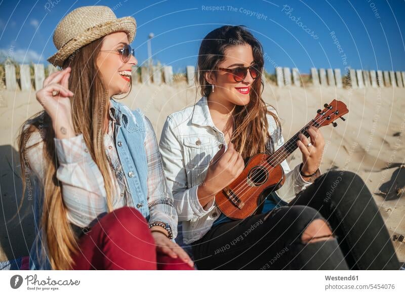 Zwei junge Frauen mit Ukulele am Strand sitzend Beach Straende Strände Beaches Freundinnen Freunde Freundschaft Kameradschaft weiblich sitzt lachen Sonnenbrille