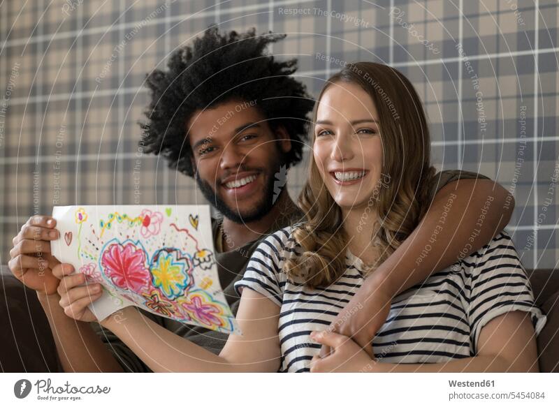 Lächelndes Paar sitzt auf der Couch und zeigt eine Kinderzeichnung Kinderzeichnungen Pärchen Paare Partnerschaft lächeln Zeichnung Zeichnungen Bild Bildnis