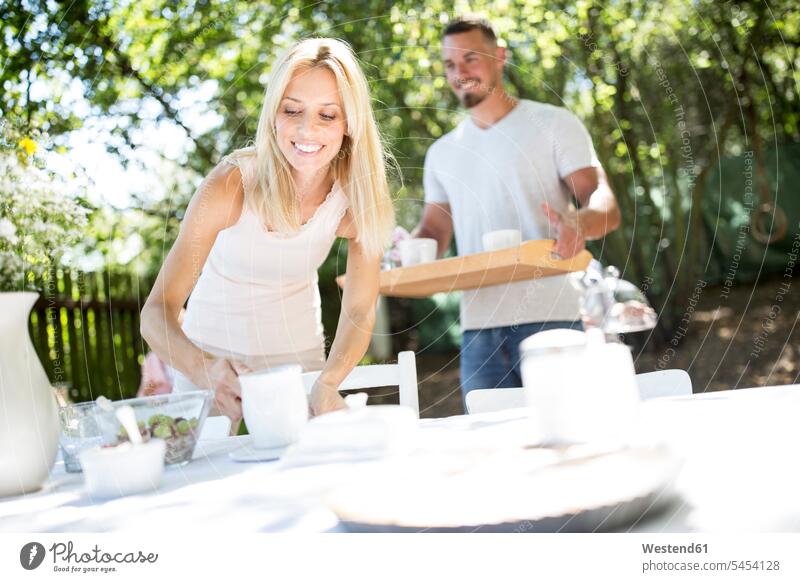 Lächelndes Paar deckt Gartentisch lächeln Pärchen Paare Partnerschaft Tisch Tische Mensch Menschen Leute People Personen bringen mitbringen Freizeitkleidung