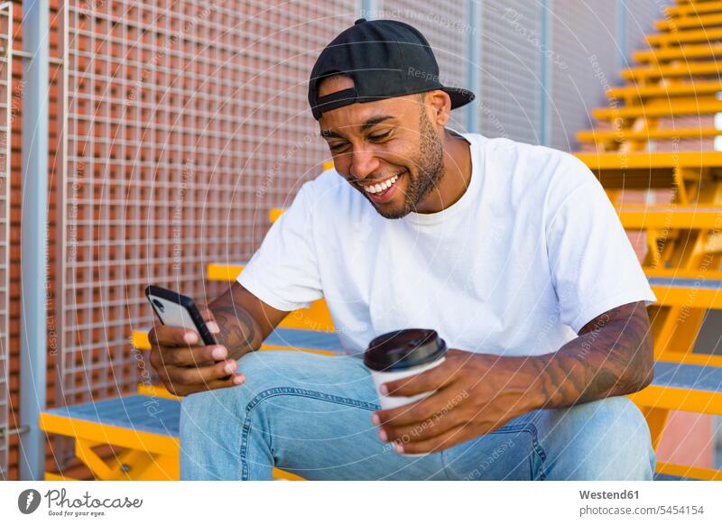 Lachender junger Mann mit Kaffee, der auf der Treppe sitzt und sich ein Smartphone ansieht lachen iPhone Smartphones Männer männlich positiv Emotion Gefühl
