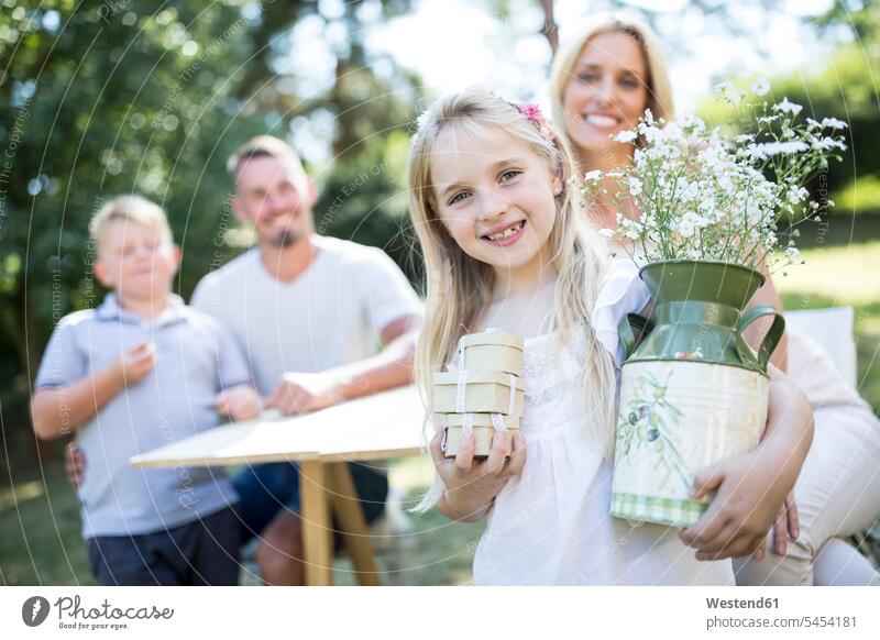 Porträt eines lächelnden Mädchens im Freien, das einen Krug und Kisten mit der Familie im Hintergrund hält Blume Blumen Blüte Krüge Kruege Pflanze Pflanzenwelt