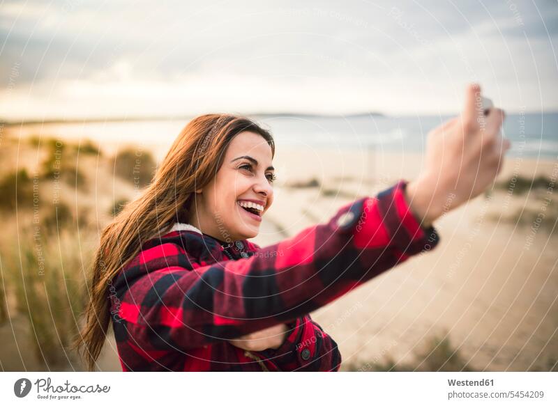 Lachende junge Frau, die ein Selfie mit ihrem Smartphone am Strand macht Selfies Beach Straende Strände Beaches weiblich Frauen Erwachsener erwachsen Mensch