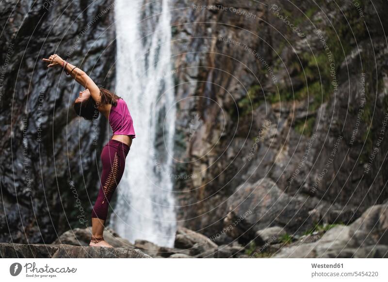 Italien, Lecco, Frau, die auf einem Felsen in der Nähe eines Wasserfalls eine Standing Backbend Yoga Pose macht Yoga-Übungen Yogauebungen Yogaübungen Jogauebung