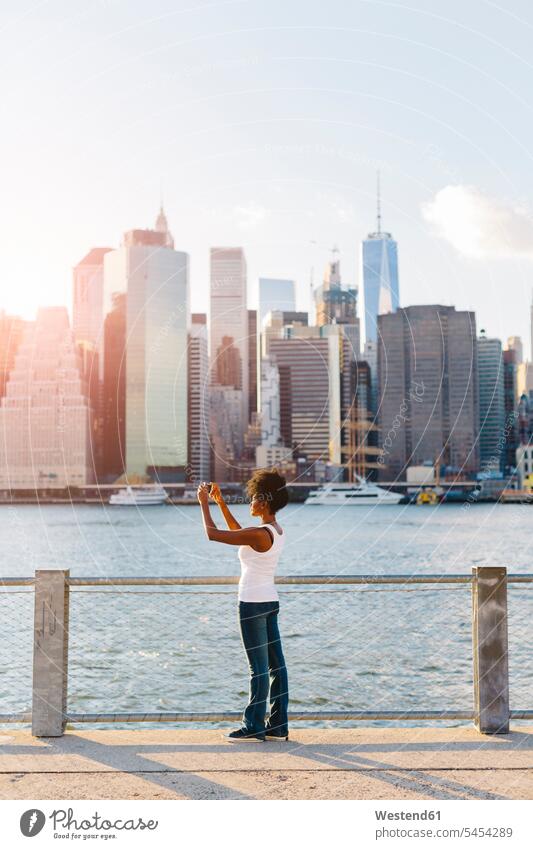USA, New York City, Brooklyn, Frau steht am Wasser und macht ein Handyfoto Skyline Skylines stehen stehend weiblich Frauen Fluss Fluesse Fluß Flüsse