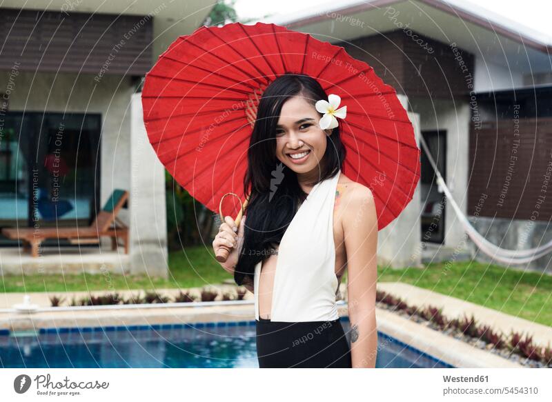 Porträt einer lächelnden Frau mit Blume im Haar, die einen roten traditionellen Regenschirm an einem Swimmingpool hält Schirm Schirme Portrait Porträts