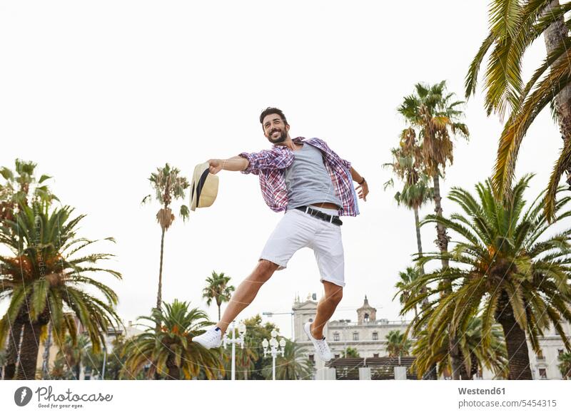 Spanien, Barcelona, glücklicher Mann springt in die Luft, umgeben von Palmen Männer männlich Portrait Porträts Portraits springen hüpfen Spaß Spass Späße