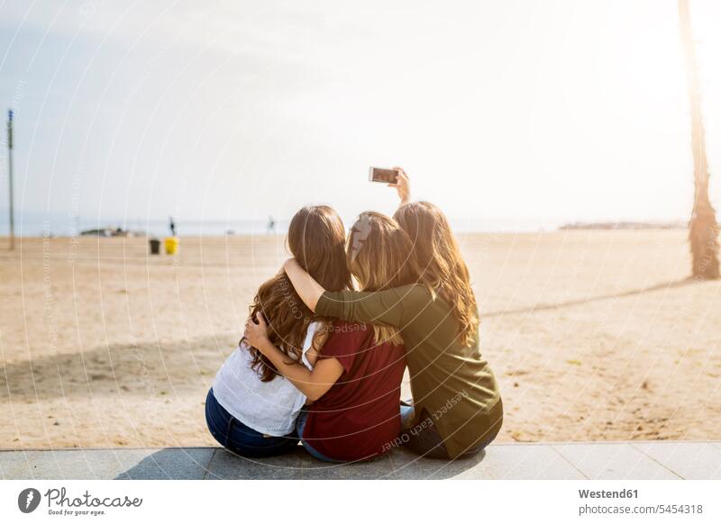 Rückansicht von drei Freundinnen, die am Strand sitzen und ein Selfie machen Handy Mobiltelefon Handies Handys Mobiltelefone Selfies Beach Straende Strände