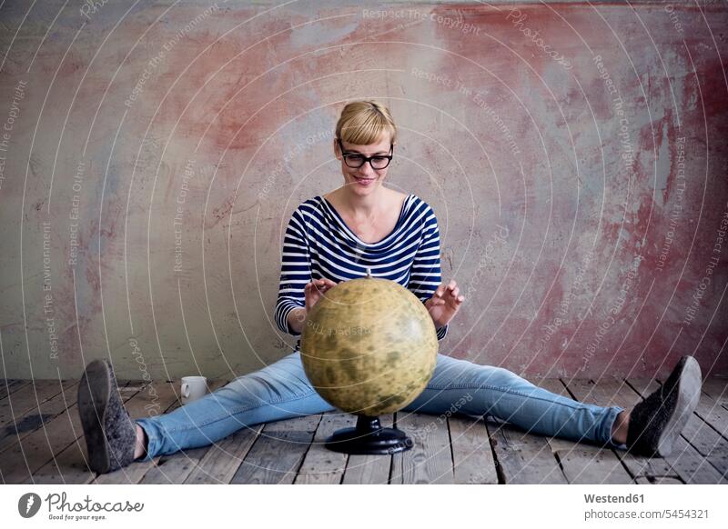 Lächelnde Frau sitzt auf Holzboden in einem unrenovierten Raum und schaut auf den Globus Weltkugel Erdkugel Globen weiblich Frauen Erwachsener erwachsen Mensch