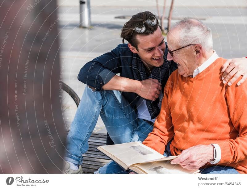 Glücklicher älterer Mann und erwachsener Enkel mit Fotoalbum im Freien Enkelsöhne Enkelsohn ansehen lächeln Bank Sitzbänke Bänke Sitzbank glücklich