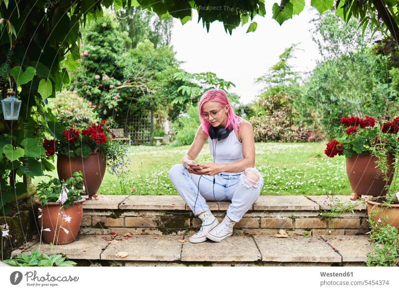 Junge Frau mit rosa Haaren trägt Kopfhörer und benutzt Mobiltelefon im Garten Kopfhoerer sitzen sitzend sitzt weiblich Frauen Handy Handies Handys Mobiltelefone