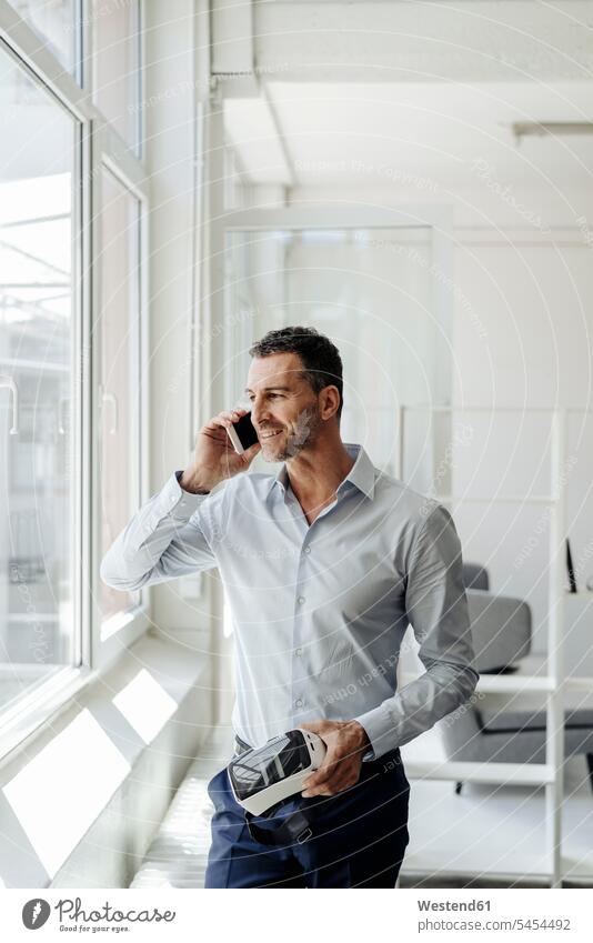 Geschäftsmann mit Handy am Fenster mit VR-Brille Mobiltelefon Handies Handys Mobiltelefone Büro Office Büros halten telefonieren anrufen Anruf telephonieren