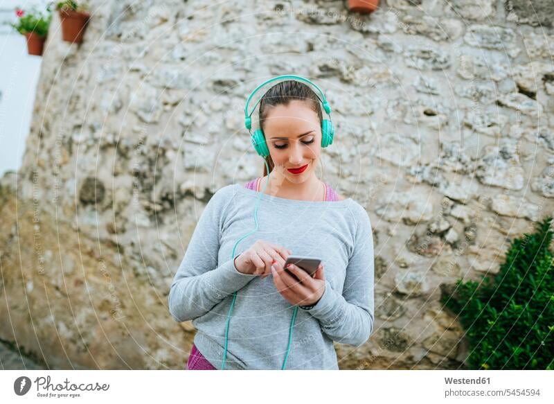 Junge Frau mit Kopfhörern sendet Nachricht mit Mobiltelefon weiblich Frauen Smartphone iPhone Smartphones lesen Lektüre SMS Textnachricht Musik hören Kopfhoerer