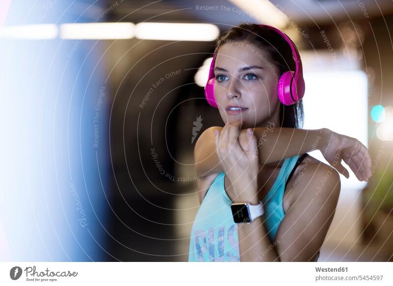 Junge Frau mit rosa Kopfhörern, die sich streckt und nachts in moderner städtischer Umgebung Musik hört trainieren Musik hören Joggerin Joggerinnen Smartwatch