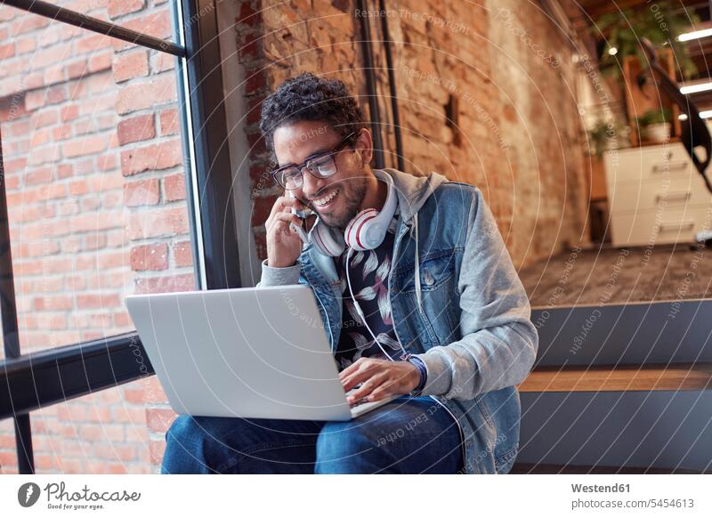 Junger Mann mit Kopfhörern und Laptop sitzt auf der Bürotreppe und telefoniert junger Mann junge Männer arbeiten Arbeit Notebook Laptops Notebooks Unternehmen