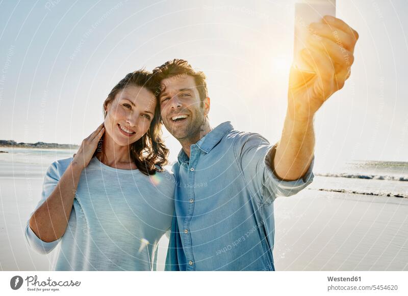 Porträt eines glücklichen Paares, das mit seinem Smartphone ein Selfie am Strand macht Beach Straende Strände Beaches Selfies Pärchen Partnerschaft Mensch