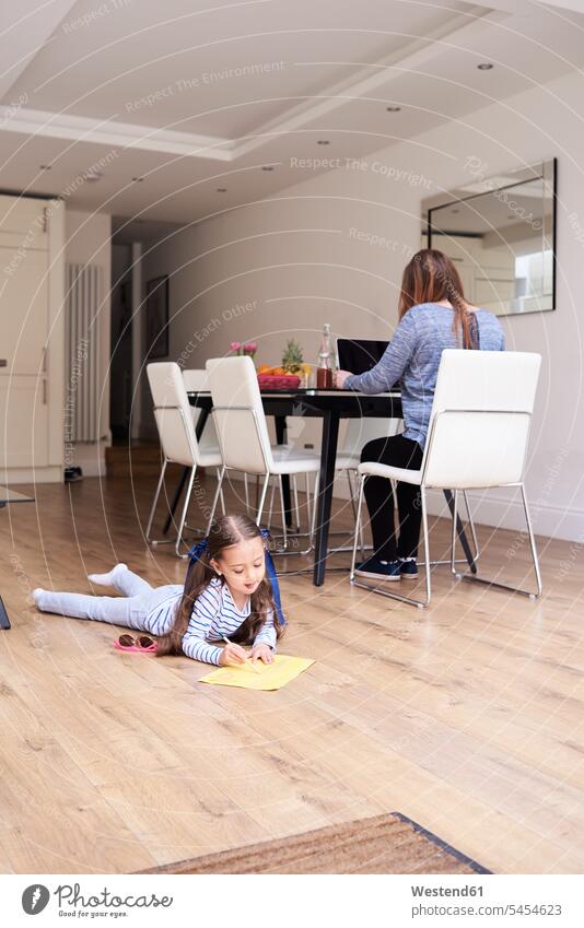 Kleines Mädchen liegt auf dem Boden und zeichnet ein Bild, während ihre Mutter im Hintergrund am Laptop arbeitet weiblich Kind Kinder Kids Mensch Menschen Leute
